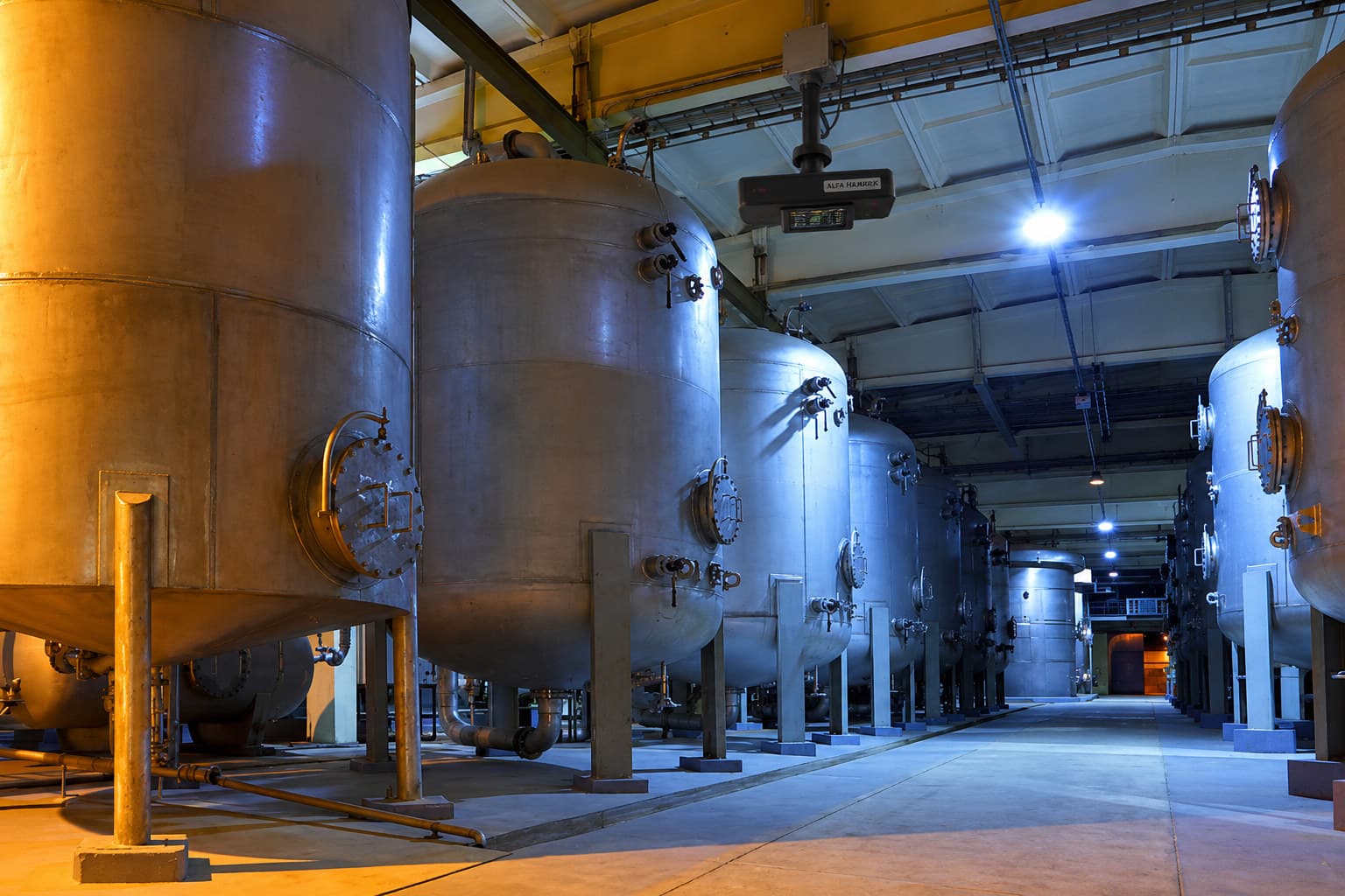A ceiling-mounted Alfa HazardX node on a drop bracket above the aisle of a process hall, with rows of large stainless steel pressure vessels and their flanged manways on either side.